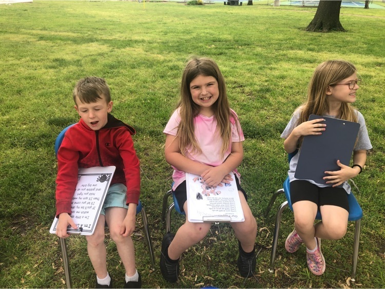 students in chairs at the park, with poems on clipboards 
