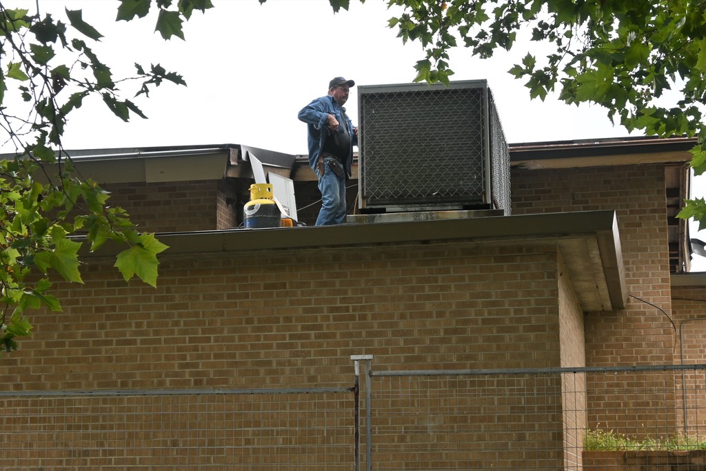 A man stands on the roof of Washington beside an air-conditioning unit  and attaches gauges to collect refrigerant.