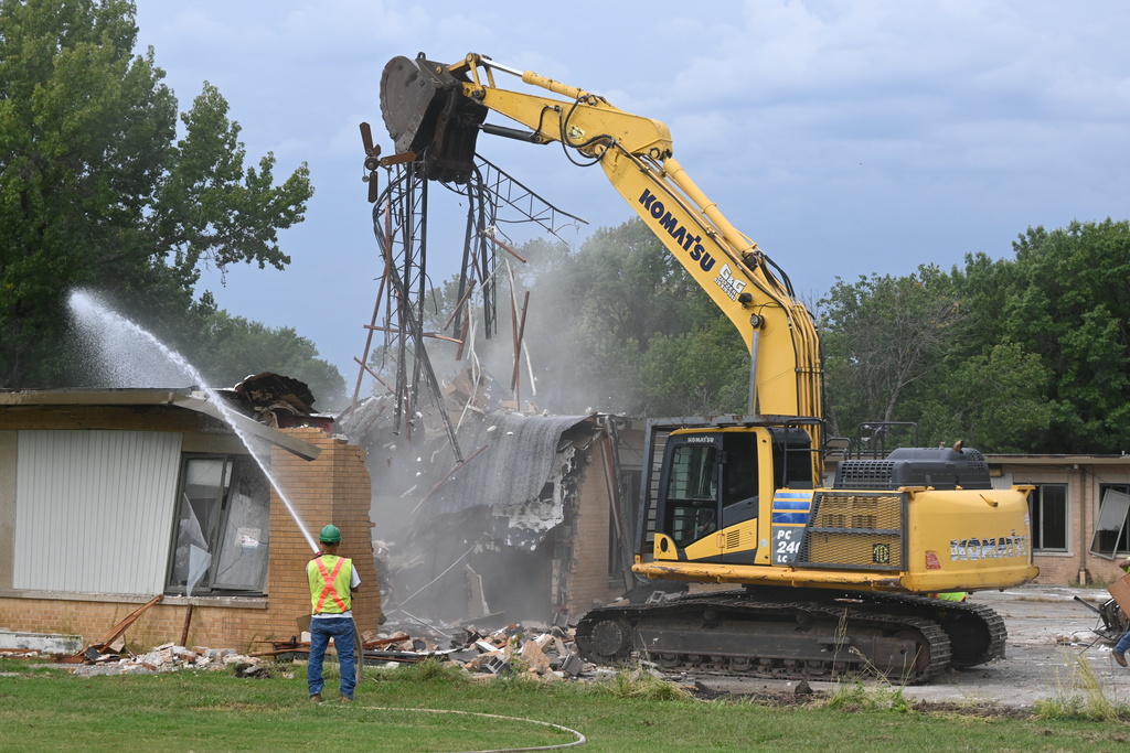 The excavator lifts metal from the roof into the air, and sticking out from the jaws is a broken ceiling fan with three blades.